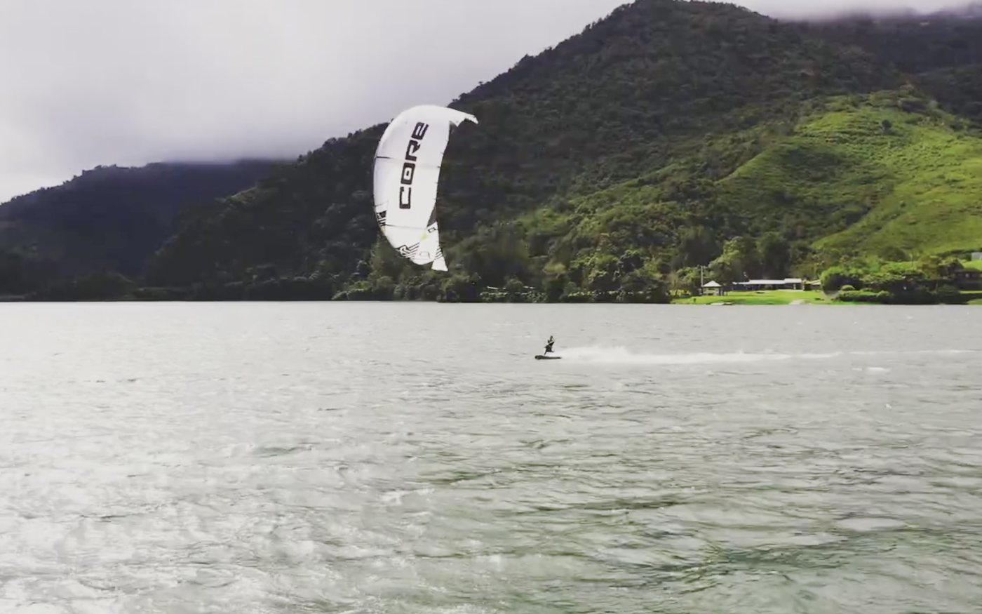 Un kitesurfista bajo una cometa blanca cruza el lago frente a lomas con nubes