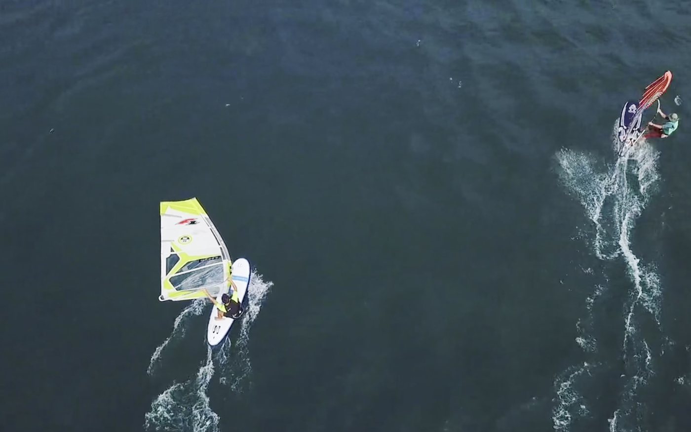 Dos windsurfistas vistos desde arriba sobre el agua oscura del lago, uno con vela amarilla