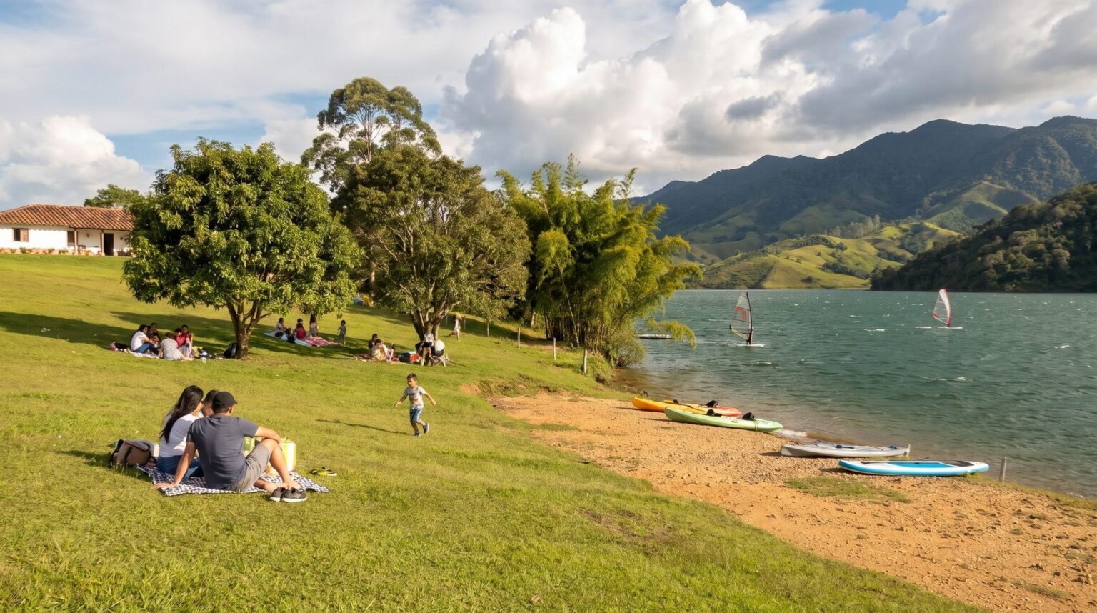 Families on picnic blankets under trees on a lawn that slopes to a small brown-sand launch beach on Lake Calima, with windsurf sails on the water.