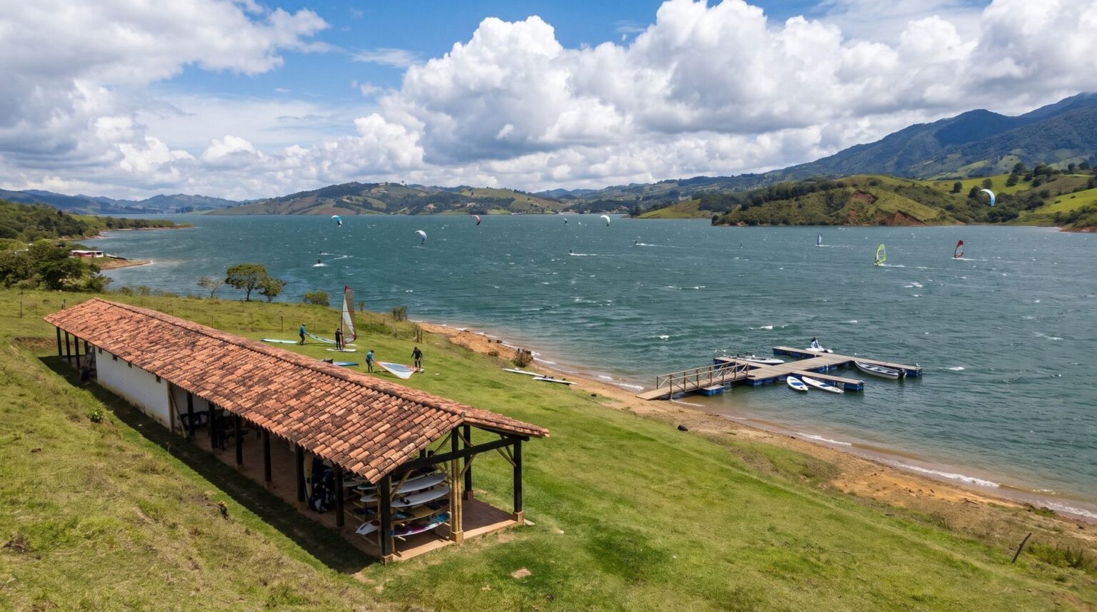 Kite and windsurf school: a low tile-roofed guadua shed set back from the shore, a floating pier on the water, and sails and kites on Lake Calima on a windy afternoon.