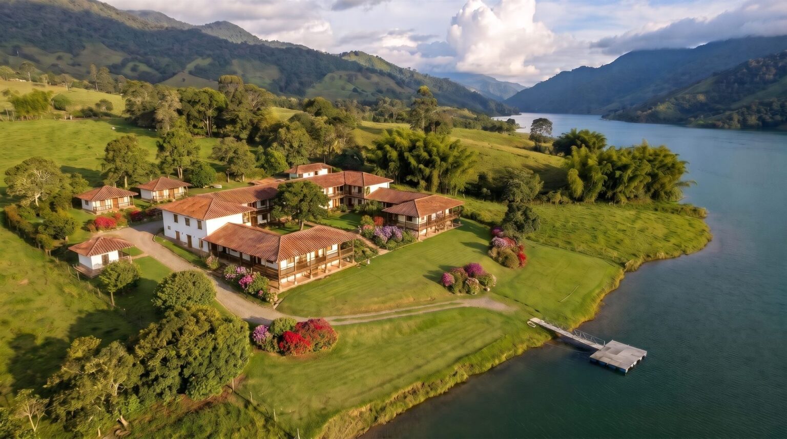 Aerial view of a country hotel of low wings with white walls and clay tile roofs around courtyards, on the slope above Lake Calima.
